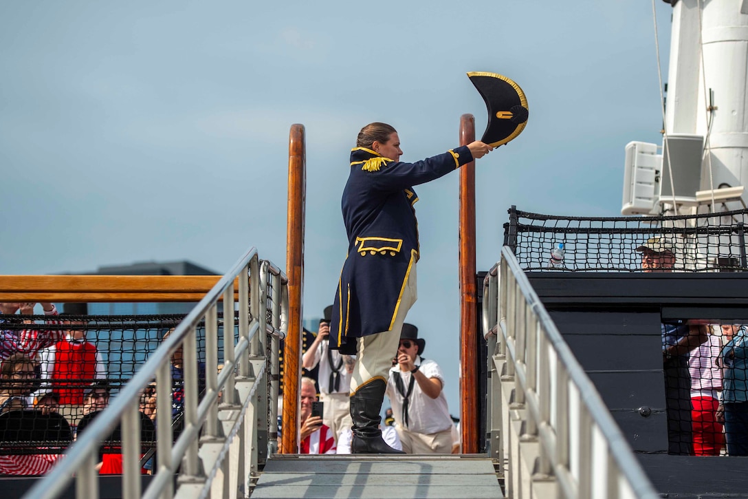 A Navy officer in period dress holds a hat in one hand while standing on the dock near a ship.