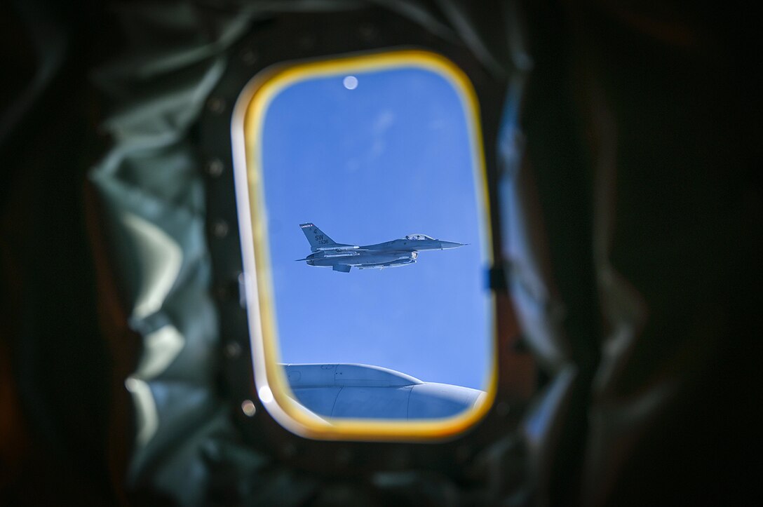 A military aircraft can be seen from the window of another aircraft, flying against a bright blue sky.