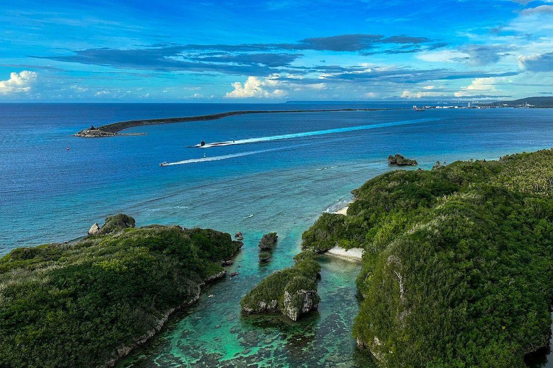 Aerial view of a Navy submarine sailing during daylight with an tree-lined shore on the right.