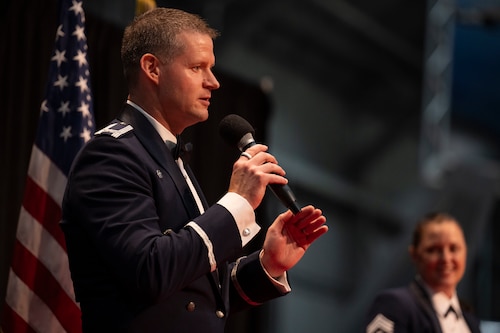 A man wearing a service uniform speaks into a microphone on stage during a graduation ceremony.