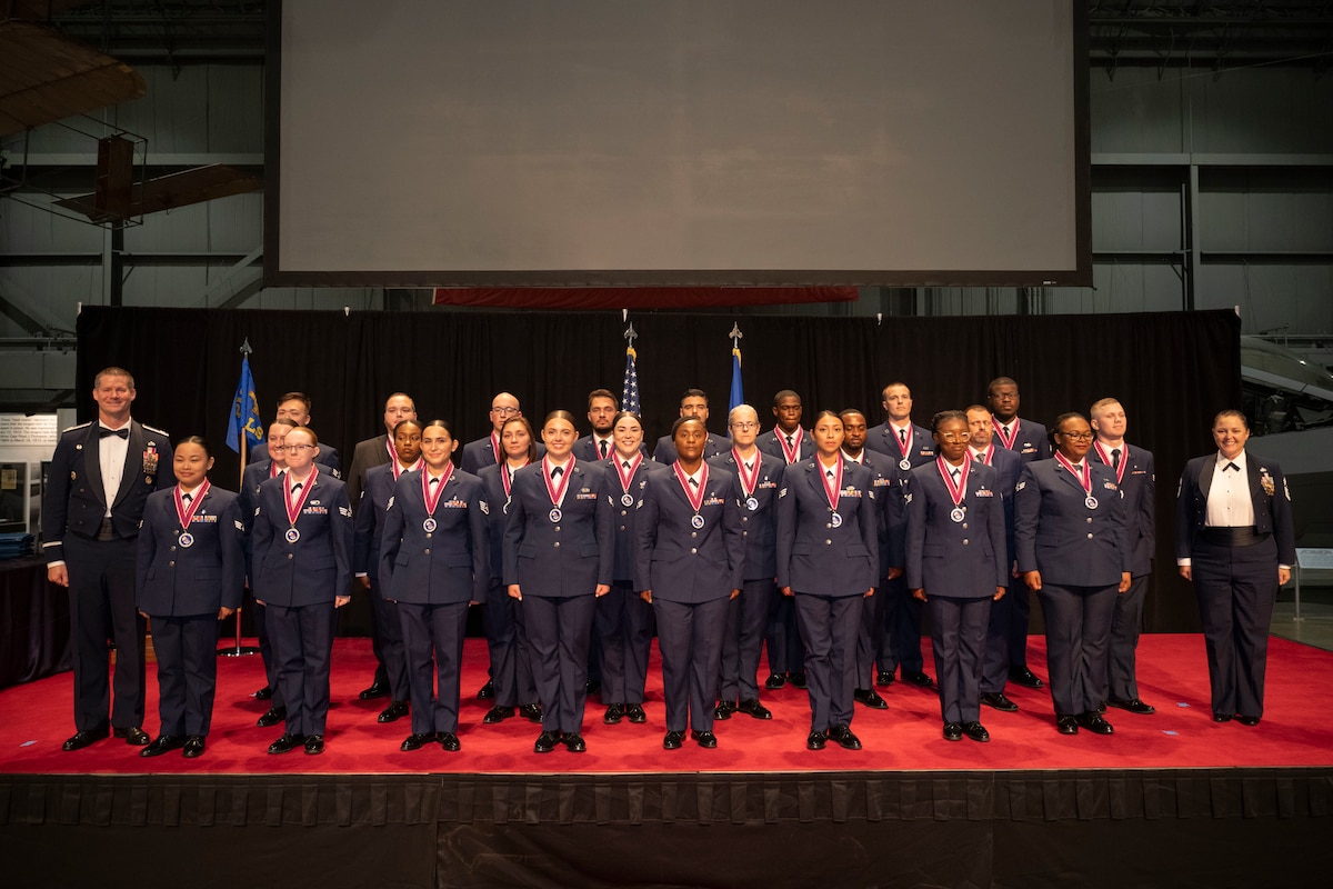 Twenty-six uniformed members stand on a red stage smiling for a group photo.