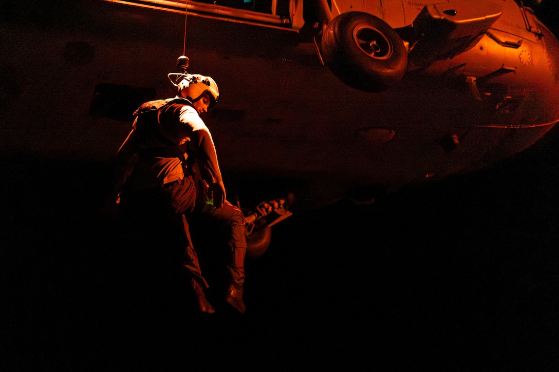 A sailor looks down while hoisted to a helicopter in the dark illuminated by a reddish light.