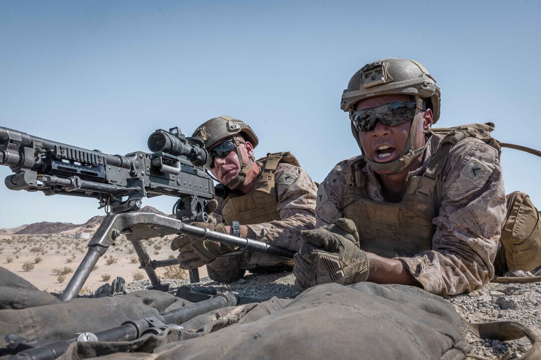 A Marine speaks while laying in a desert-like area next to another Marine aiming a weapon.