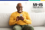 Elderly man wearing glasses sits on couch and looks at smartphone.
