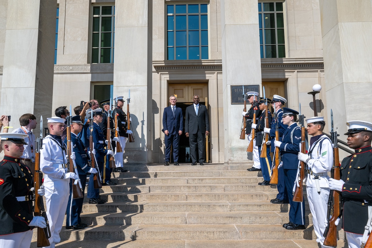 Two men pose on the Pentagon steps surrounded by service members standing in formation carrying ceremonial rifles.