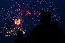 People are silhouetted as they watch fireworks explode in a dark sky.
