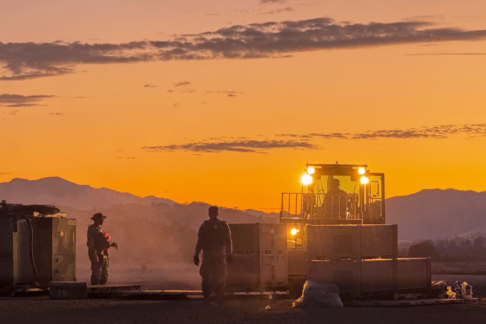 An airman operates a construction vehicle as two fellow airmen stand near boxes in a desert-like area with mountains in the background under an orangish sky.