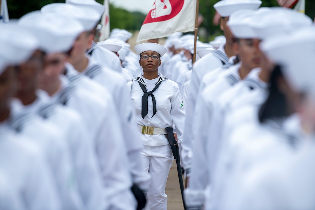 A Navy recruit stands in the center, flanked by rows of other recruits shown out of focus in the photo.