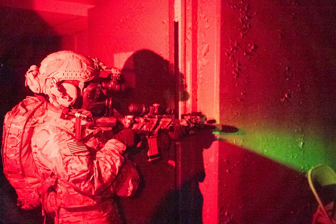 A soldier wearing protective gear aims a weapon reflecting a green light off of a concrete wall illuminated by red light.