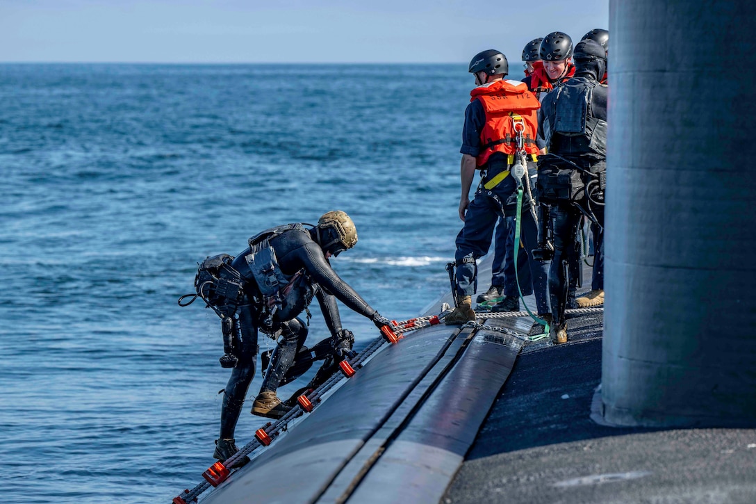 A sailor in a wet suit climbs onto a submarine from a body of water as a group of fellow sailors stand to the right.