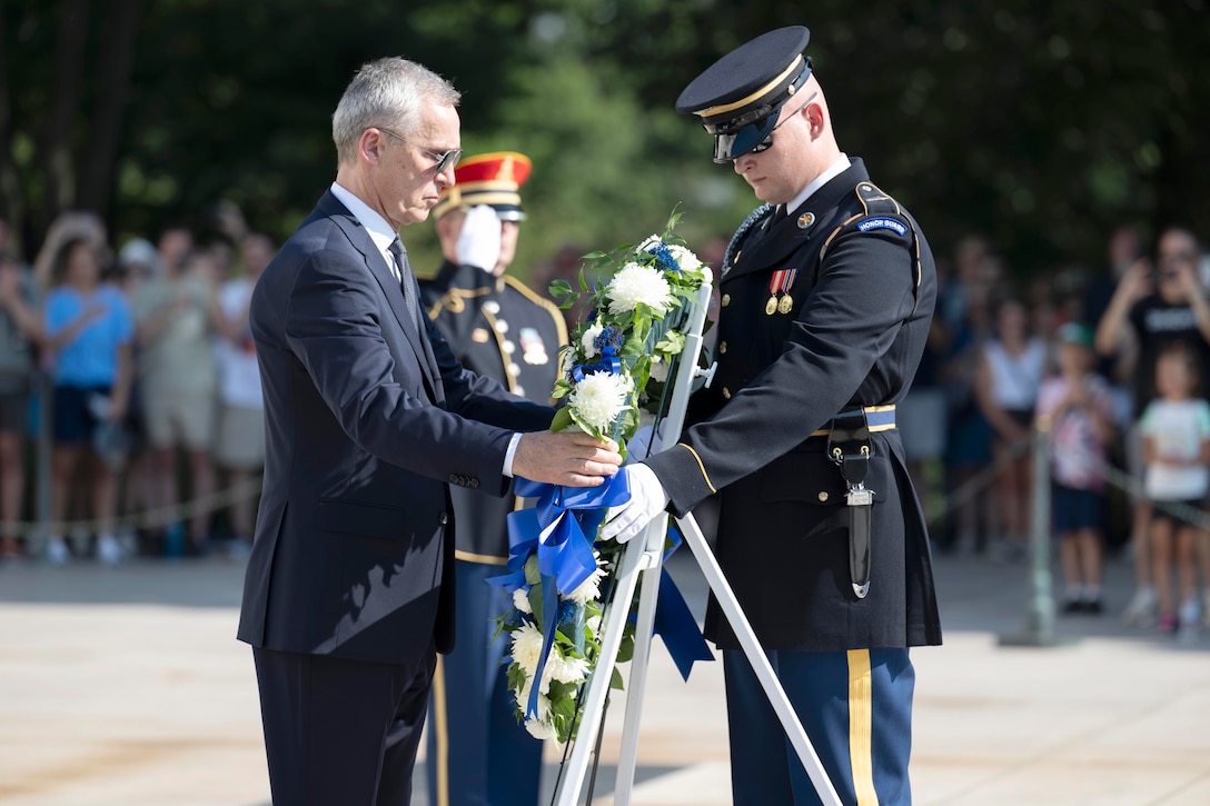 A service member and a person hold a wreath from opposite ends as a crowd watches in a blurred background.