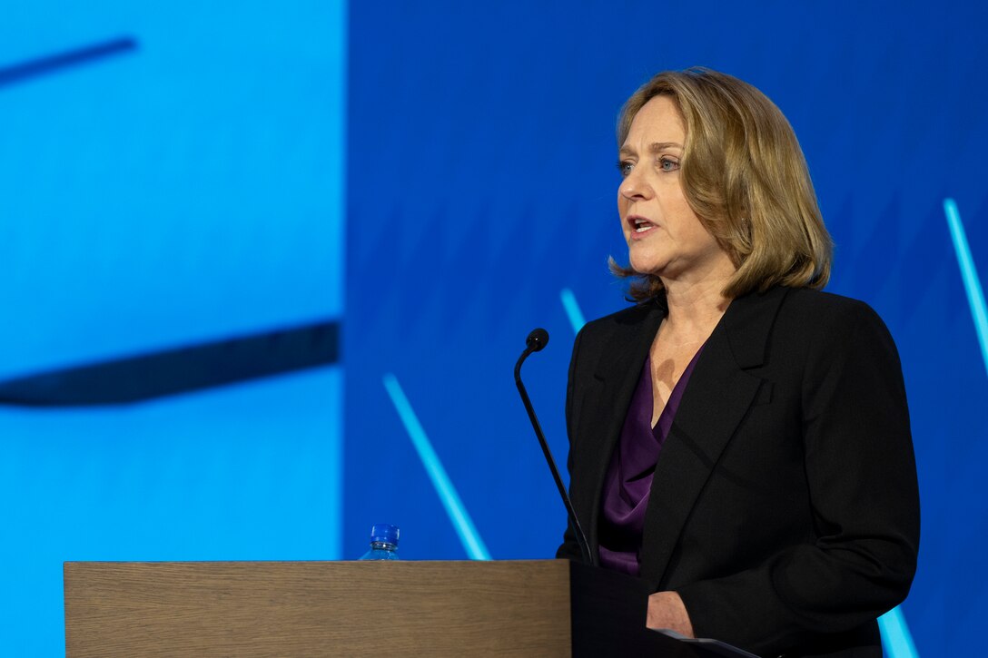 Deputy Defense Secretary Kathleen Hicks speaks into a microphone at a lectern, with a blue background behind her.
