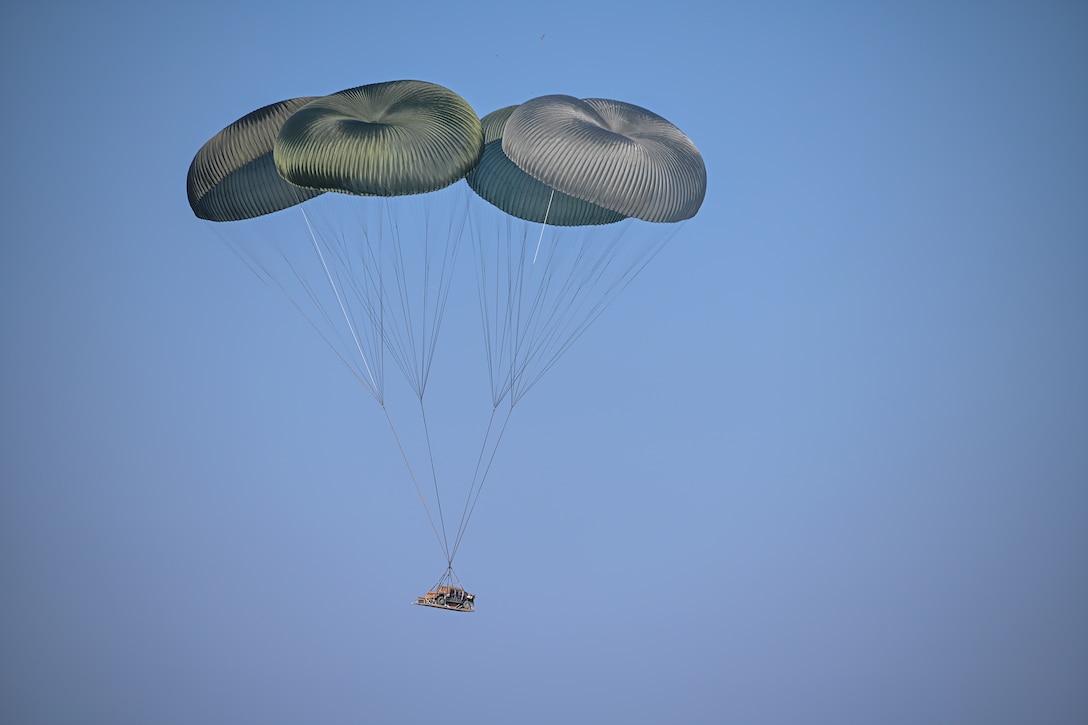 A Humvee attached to four parachutes with open canopies descends in a blue sky.