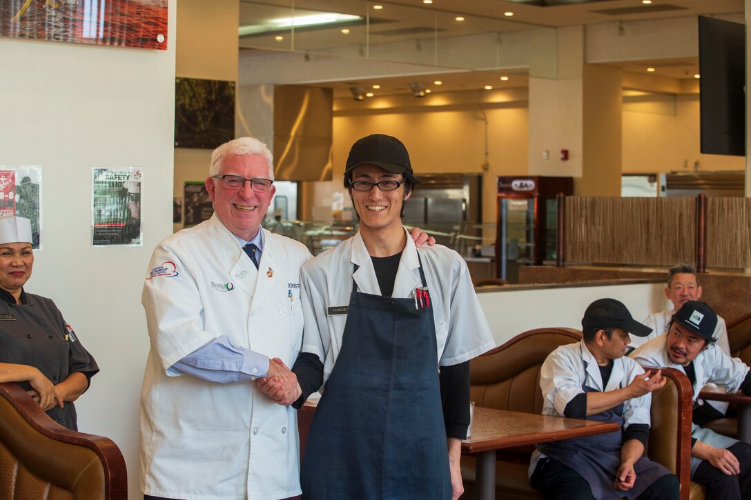 Mike Johnston, left, a member of the National Restaurant Association Education Foundation, gives a challenge coin to Takashi Yamada, the chief cook of the Flight Line Mess Hall at Marine Corps Air Station Iwakuni, Japan, for outstanding work while taking part in the Maj. Gen. William Pendleton Thompson (W.P.T.) Hill memorial awards program, Jan. 29, 2024. The W.P.T. Hill awards program aims to acknowledge outstanding performance within Marine Corps food service operations, including determining and recognizing the best mess halls of the year.