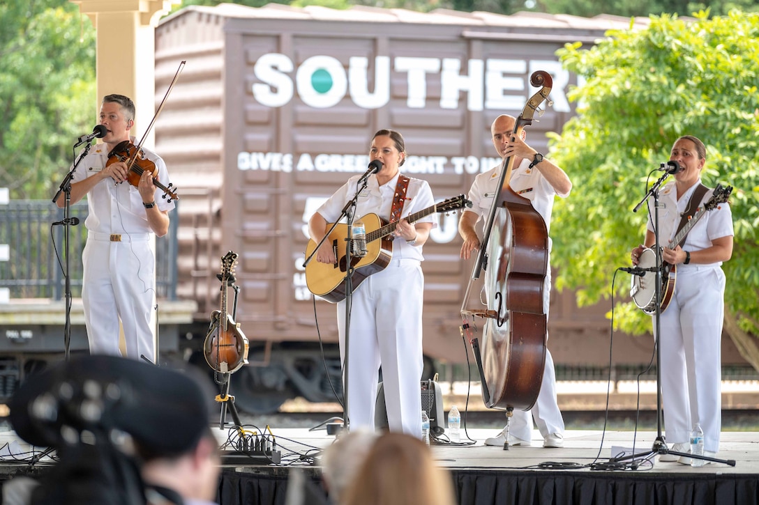 Four sailors in formal uniforms stand on a stage and play various instruments.