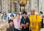 A family of four, a samll boy, a mom, a young teen boy and a dad stand with arms around each other looking at the camera while standing inside a classical Italian church