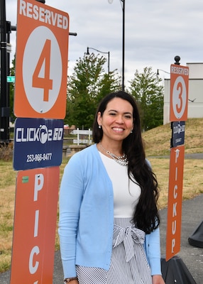 A woman in a blue sweater stands outside next to a sign that instructs people who used the click 2 go app to park there to get their order.