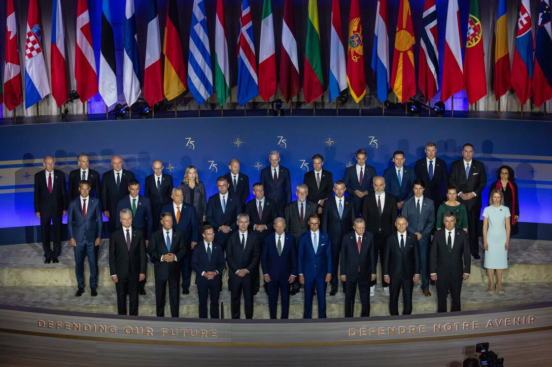 Men and women wearing business attire stand on a stage before a row of flags.