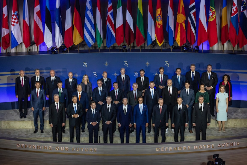 Men and women wearing business attire stand on a stage before a row of flags.