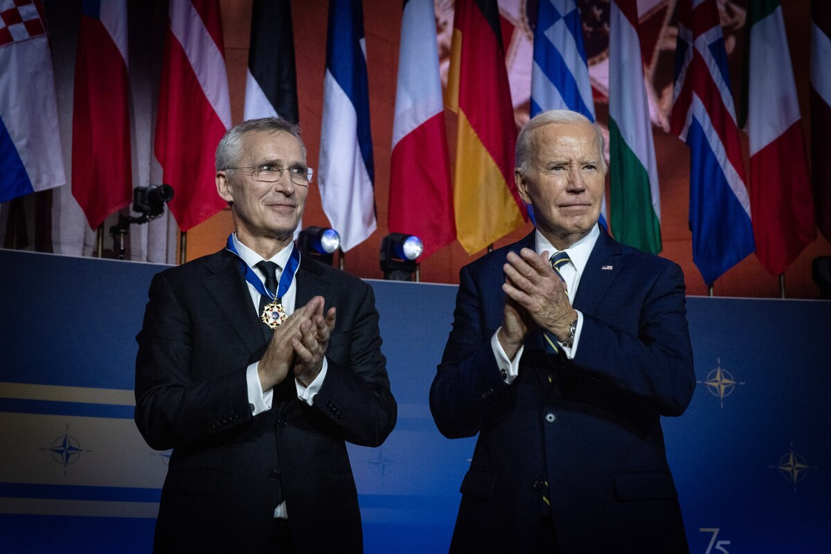 Two men in business suits, one wearing a medal, clap while standing on a stage before a row of flags.