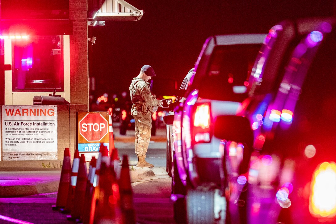 Cars line up outside of a gate as an airman stands outside of a small structure checking a driver's identification.