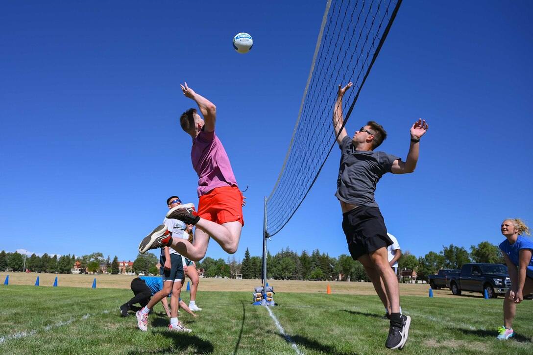 A service member in athletic clothing jumps to strike a volleyball over a net as others participate in the game.