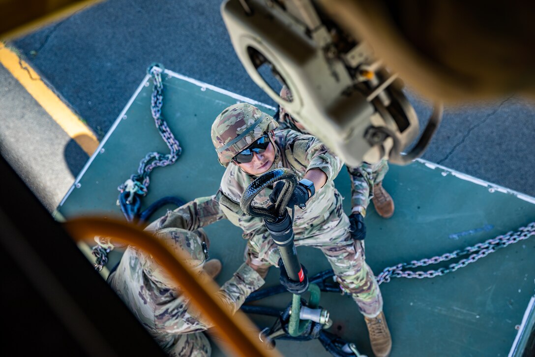 A photo of a soldier handling a sling-load below as two others provide support.