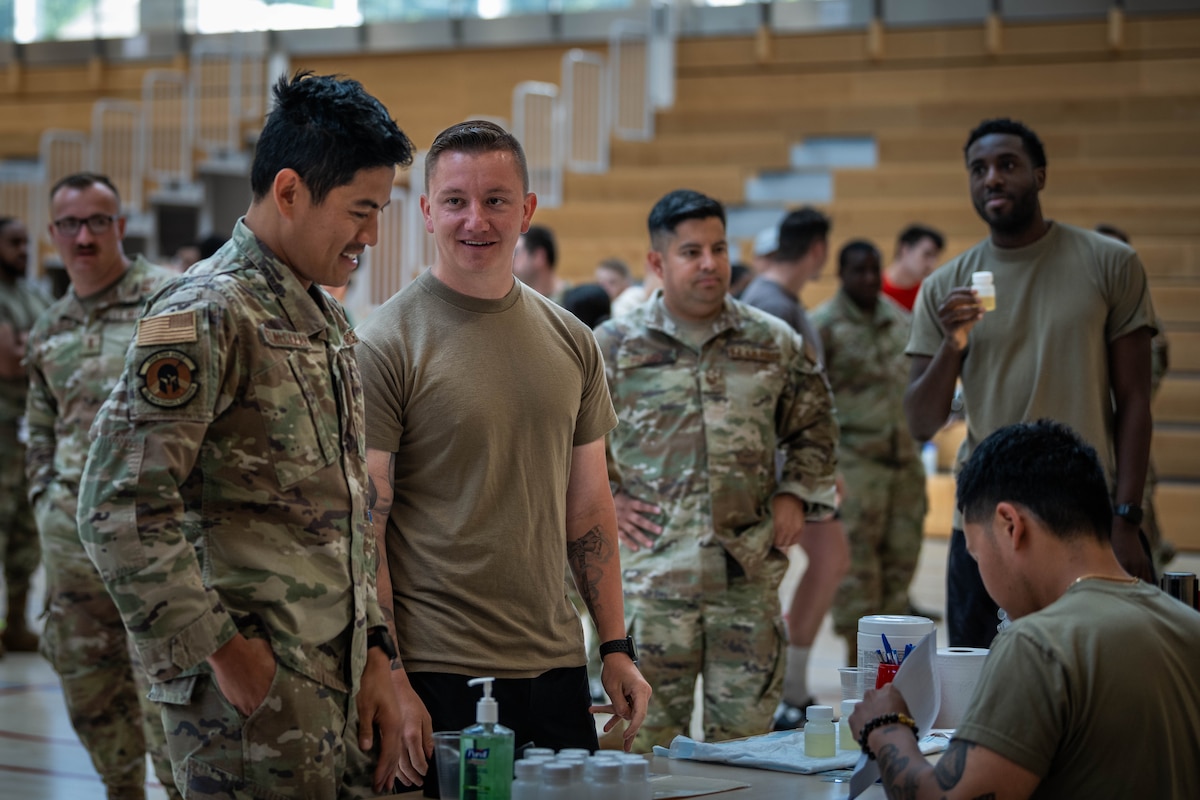 Airmen stand in from of a table while a urinalysis monitor reviews paperwork inside a gym.