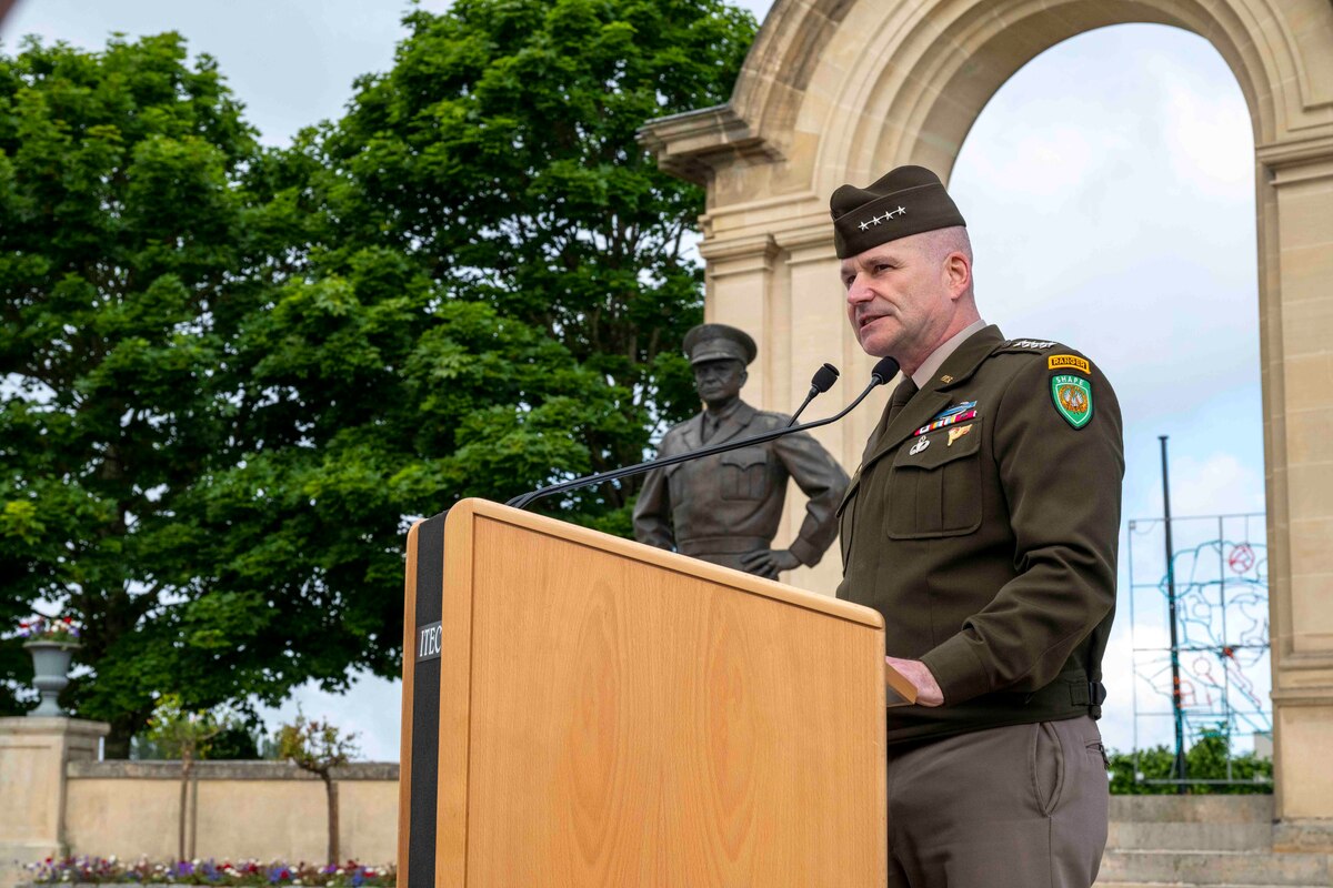 A man in an army uniform is standing at a lectern in front of an archway and a statue of another man in an army uniform.