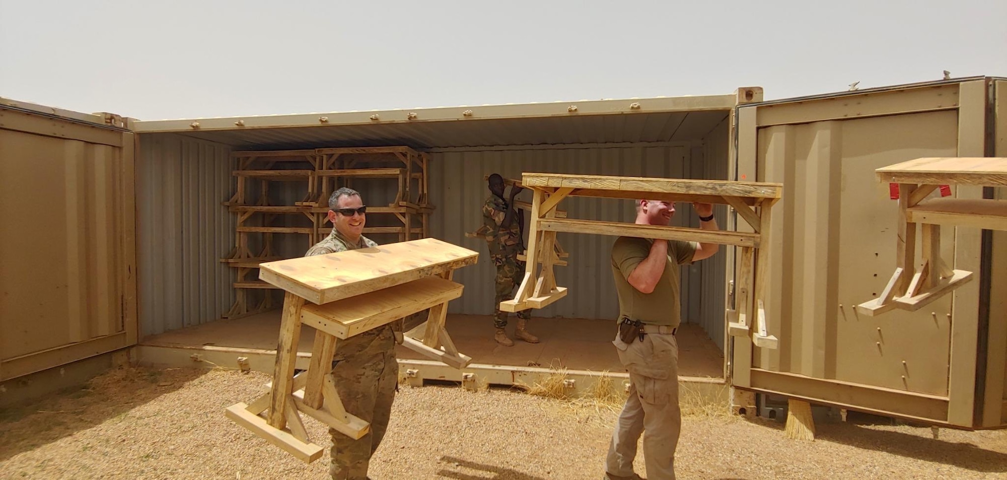 Uniformed servicemembers carry wooden desks from a storage container in a desert environment
