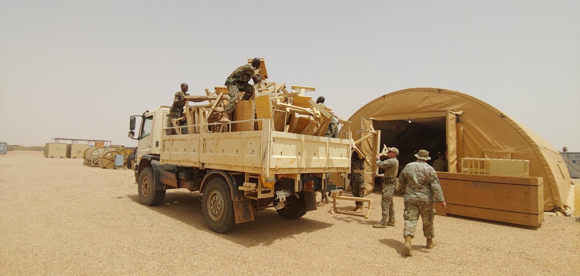 Military members in uniform load wooden desks onto a truck in a desert environment