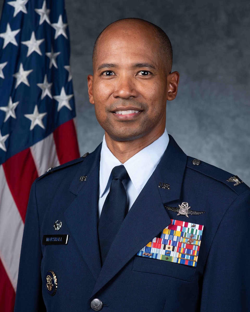 An Air Force Colonel in dress blues looks forward in front of an American flag and a grey background.