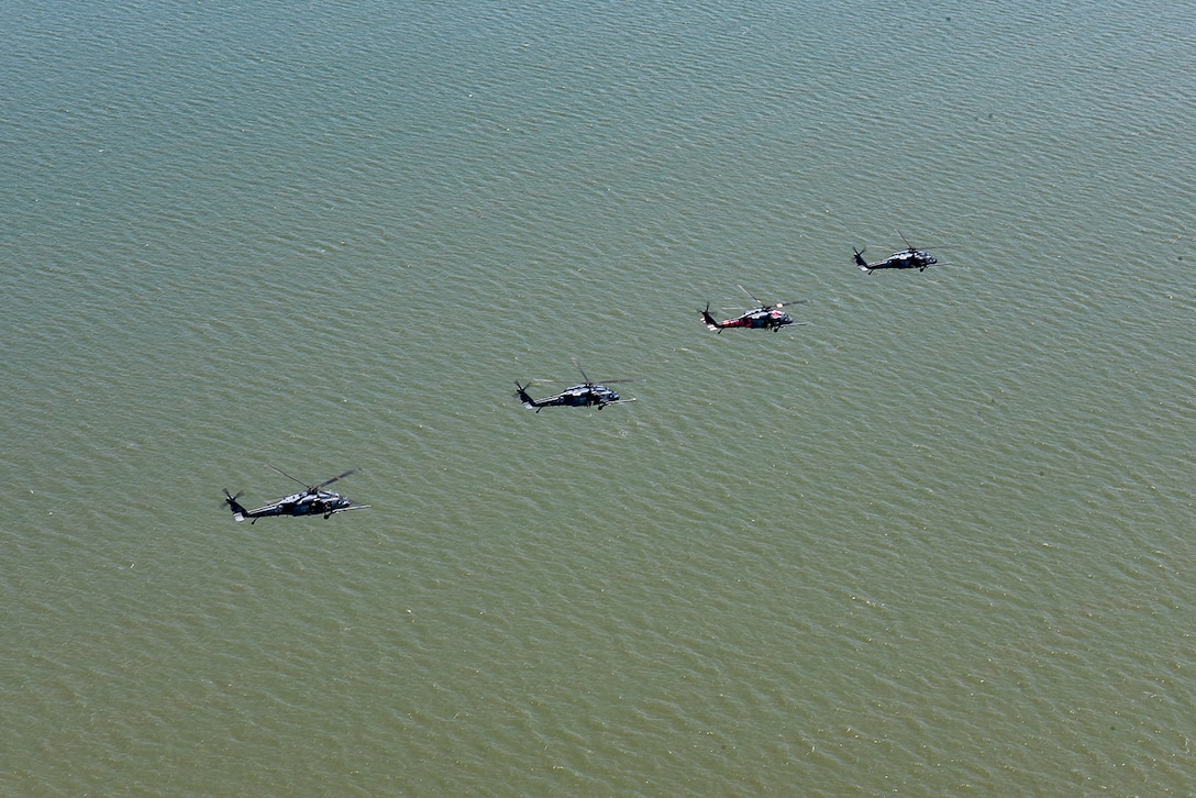 Four helicopters fly over a body of water during daylight.