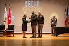 man wearing U.S. Army uniform accepts a certificate from a man and woman on stage.