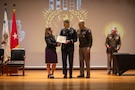 man wearing U.S. Army uniform accepts a certificate presented from a man and a woman.