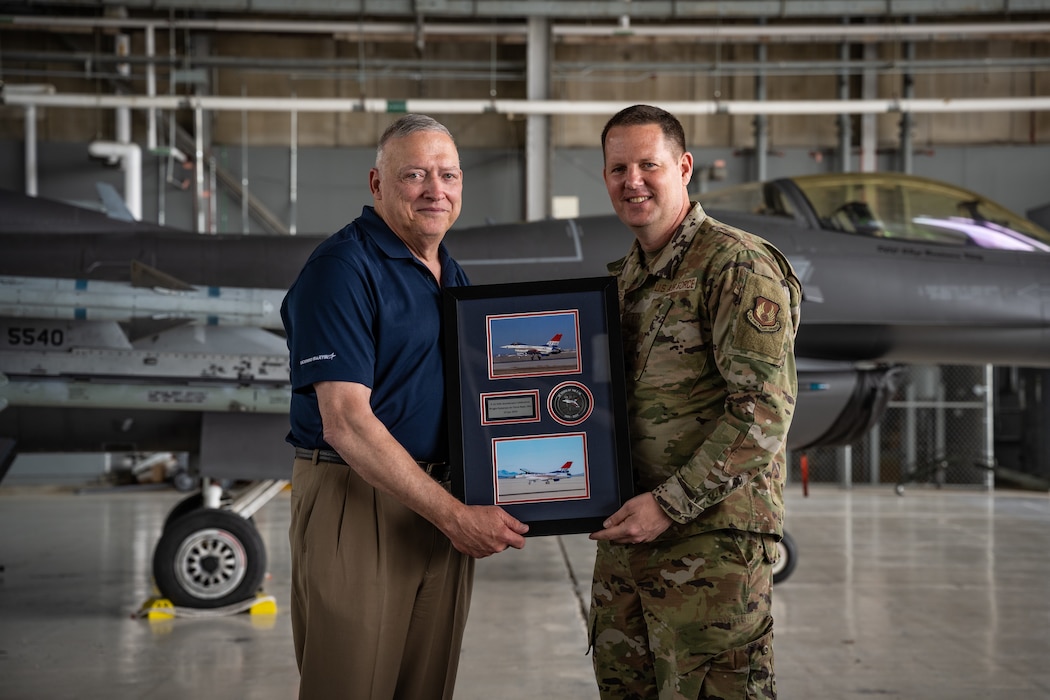 Retired Gen. North and Brig. Gen. Voorheis pose for a photo while holding a plaque with photos of a red, white, and blue F-16 Fighting Falcon