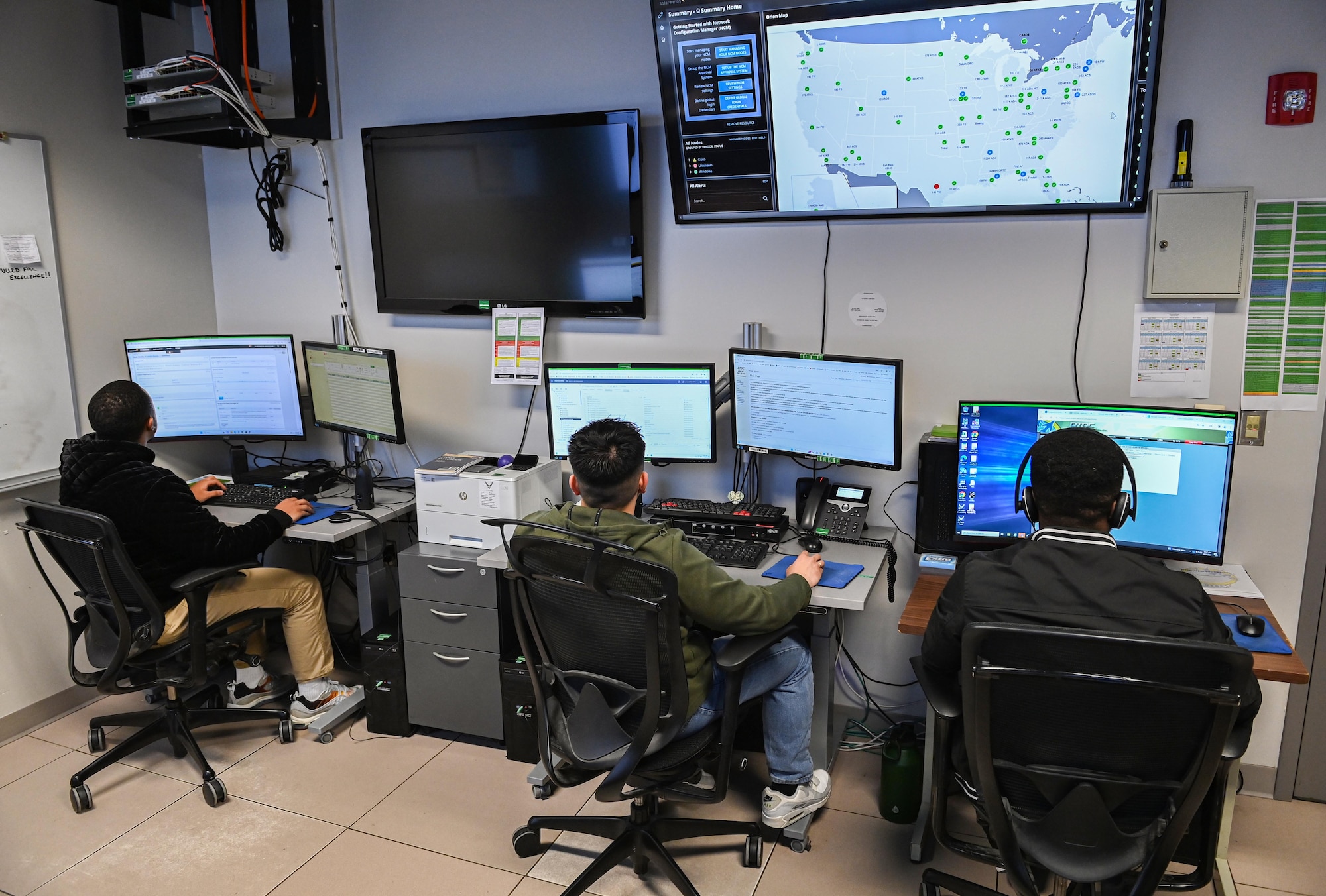 A group of civilian contractors sit facing computer screens with headsets on.