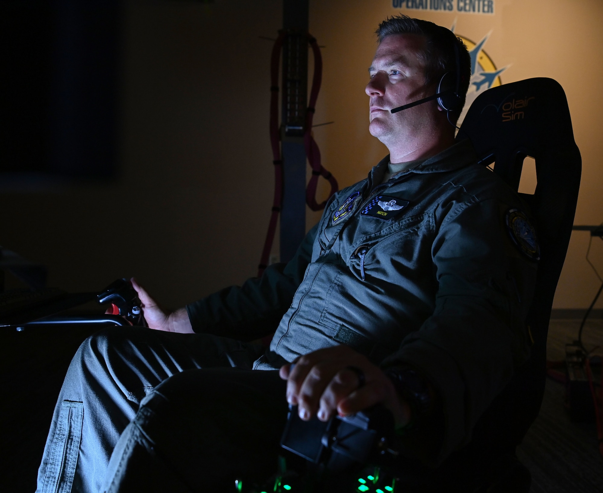 Pilot sits in a dark room at the controls of a mock flight seat with headset on.