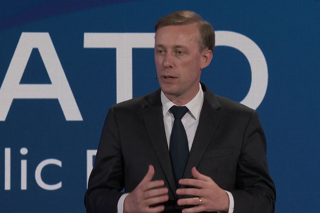 A person in business attire speaks with a blue wall in the background.