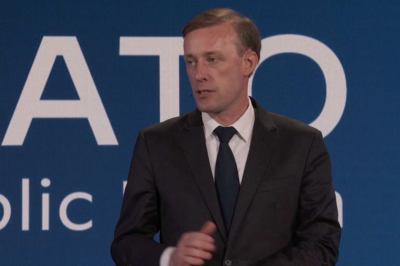 A person in business attire speaks with a blue wall in the background.