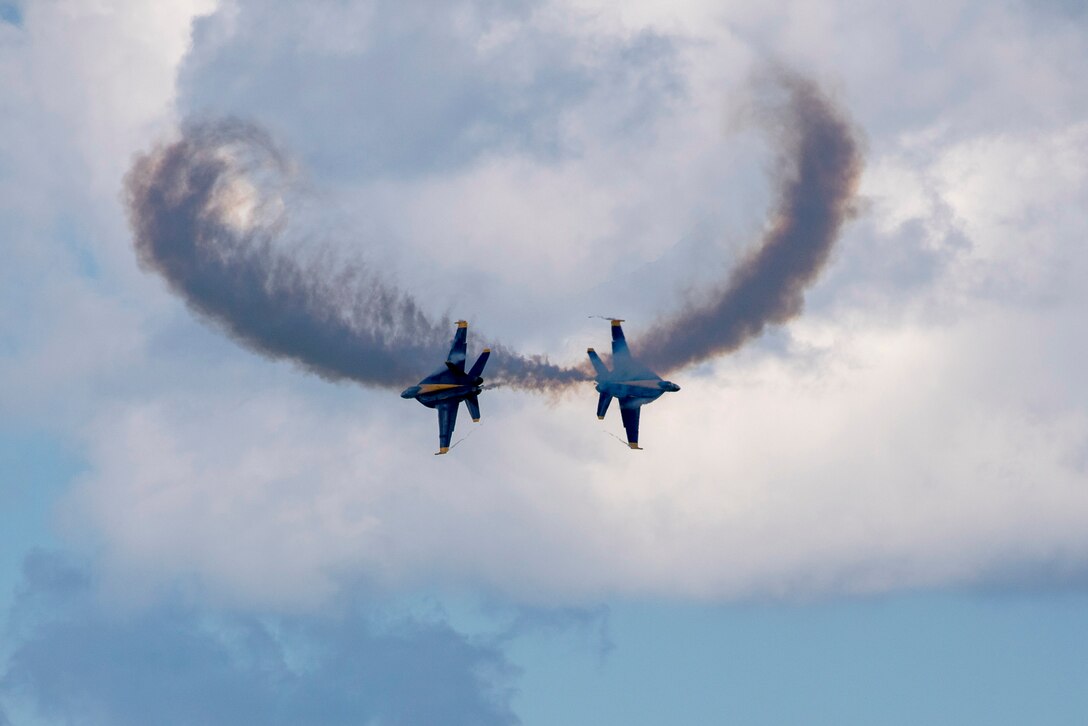 Two aircraft fly in opposite directions leaving clouds of smoke with clouds in the background.