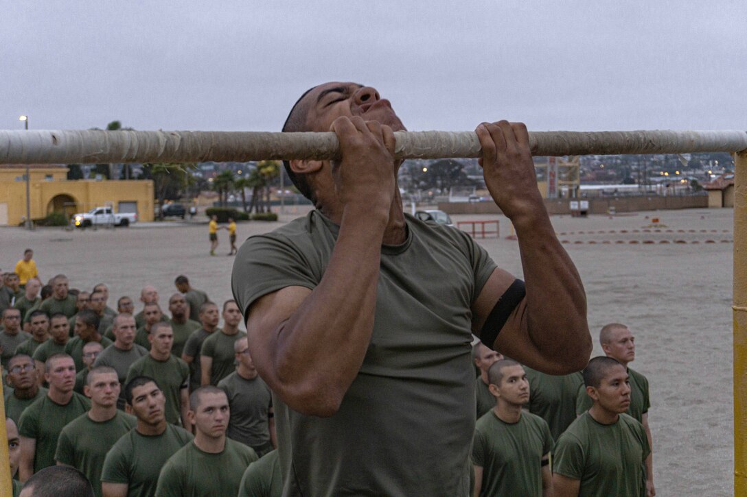 A Marine Corps recruit conducts pullups as dozens of fellow recruits stand in formation and watch from the background.