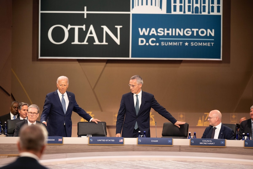 Two men in business suits grasp the back of chairs arranged along a table with microphones.