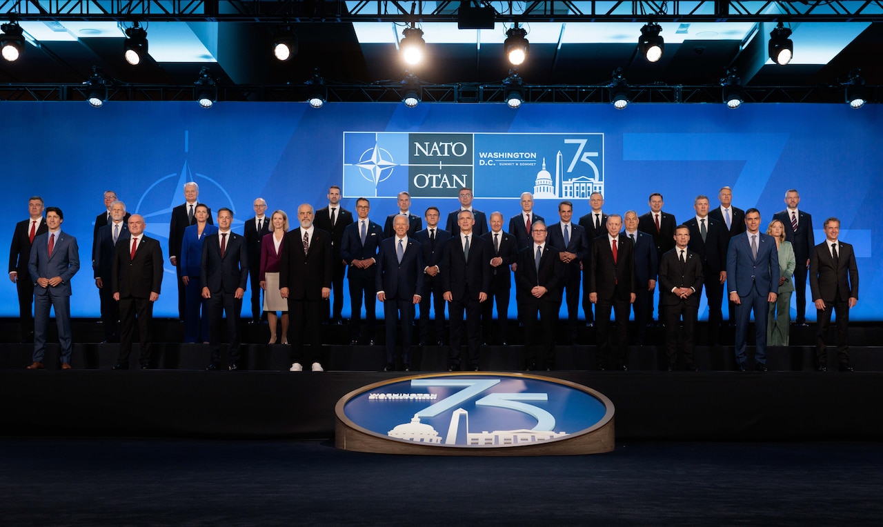 Men and women wearing business attire stand on a stage before a sign that reads “NATO.”