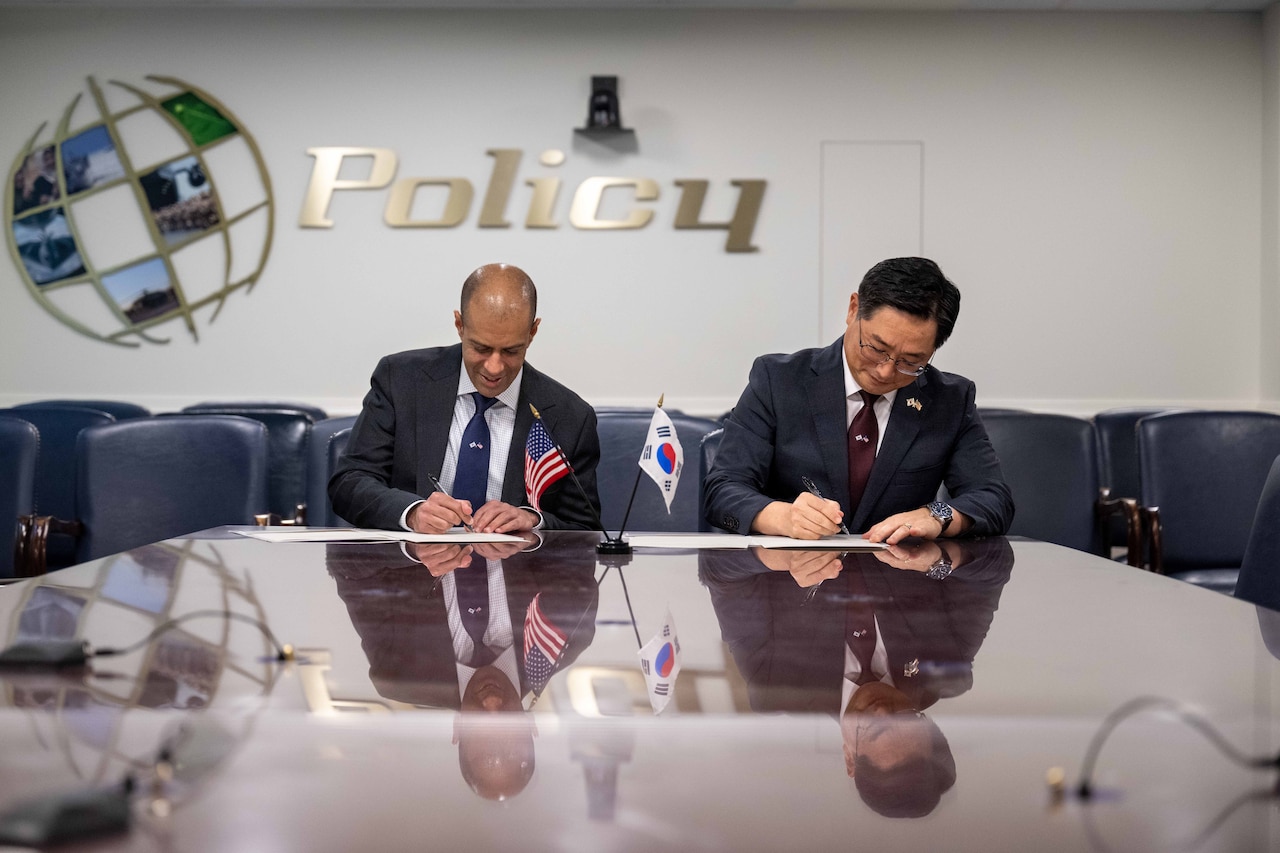 Two men wearing business suits sign a document while seated at a table displaying US. and South Korean flags.