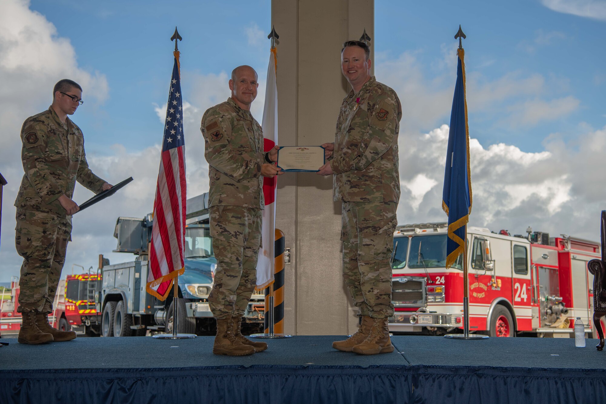 military members stand on a stage holding a medal certificate