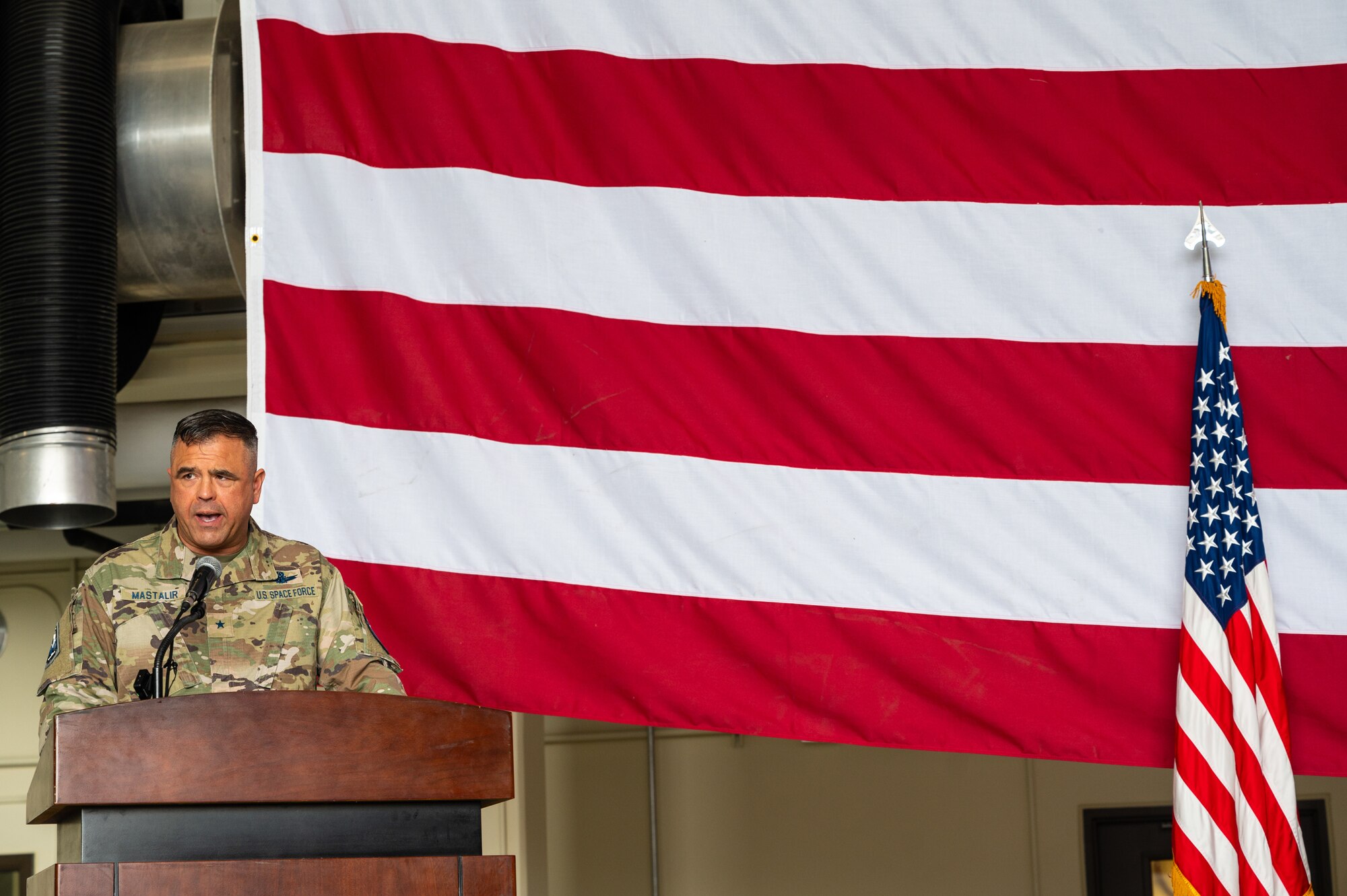 A uniformed man in camouflage stands behind a wooden podium in left corner of a photo, with a large U.S. flag hanging horizontally behind him and the U.S. flag hanging on a small post upright on the right side of the photo.
