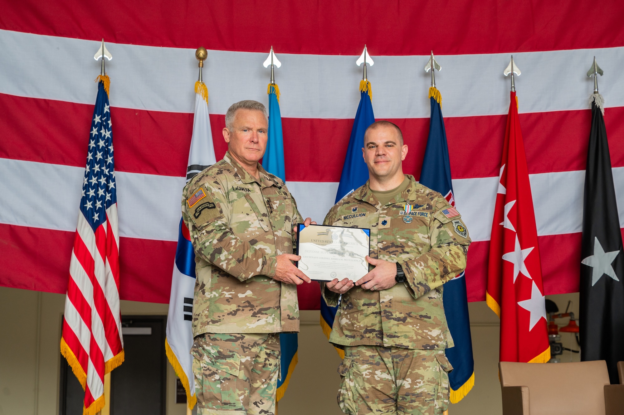 Two men in camouflage uniforms stand together displaying a certificate between them in front of a row of military flags, with a large U.S. flag hanging horizontally behind them as a backdrop.