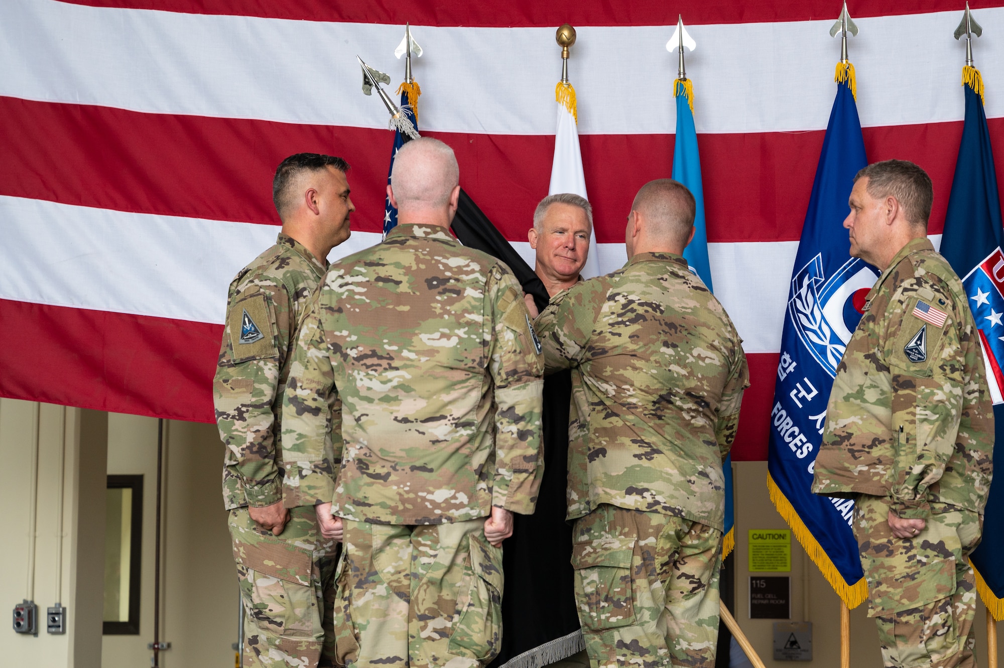 Four uniformed men stand in a diamond formation, with one on the right handing a black flag with gray tassels to one in the back, with a U.S. flag in the back-drop and a fifth uniformed man standing at attention to the right of the formation.