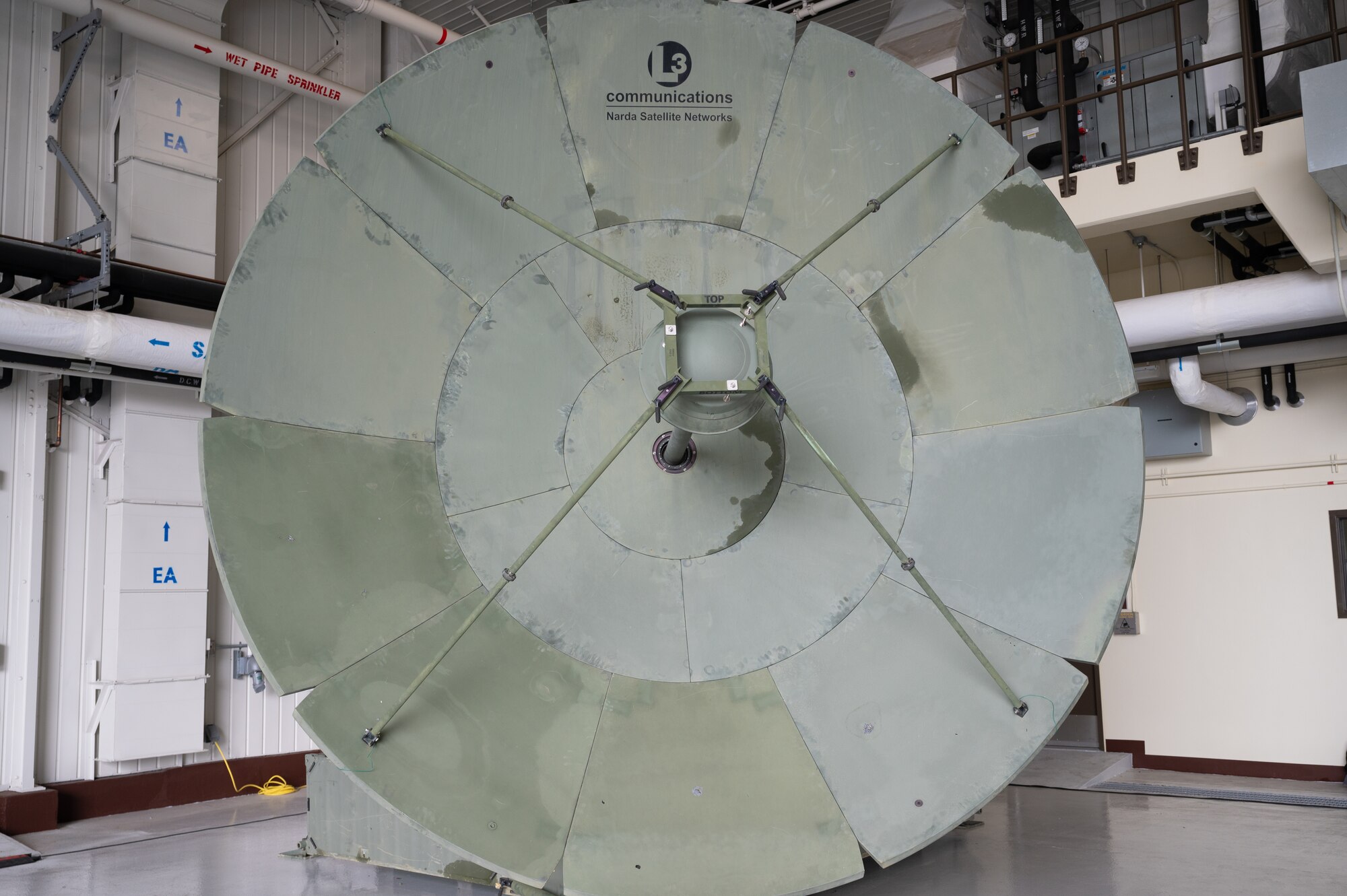 A large gray satellit dish faces the camera in the corner of a large hangar, with white wall behind it.